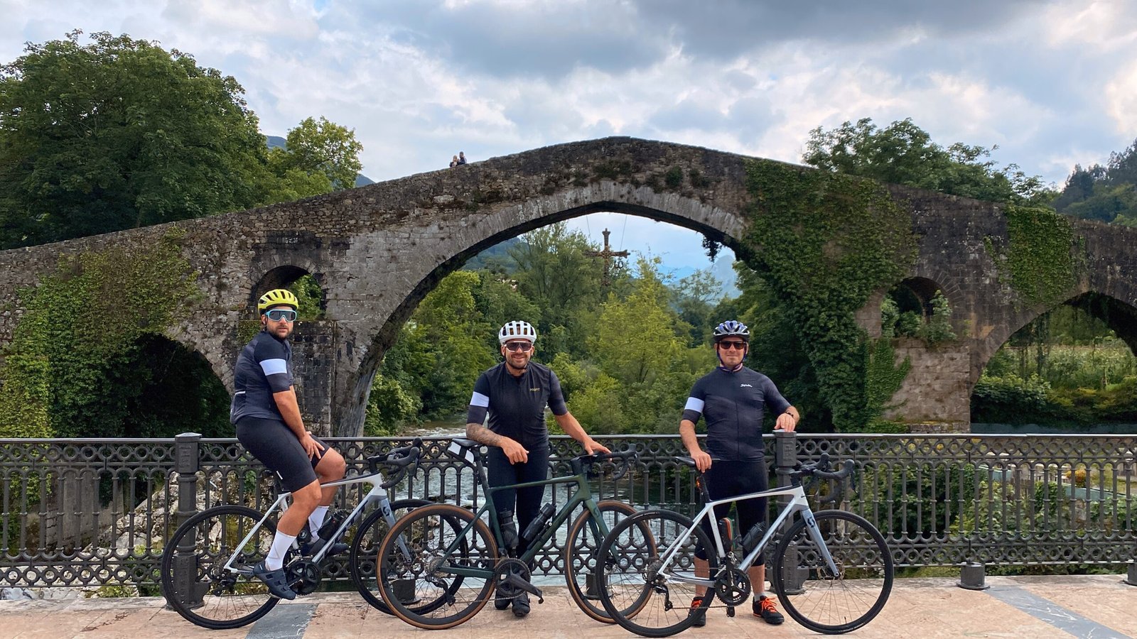 Three men with bikes posing in front of a weathered bridge in Cangas de Onis