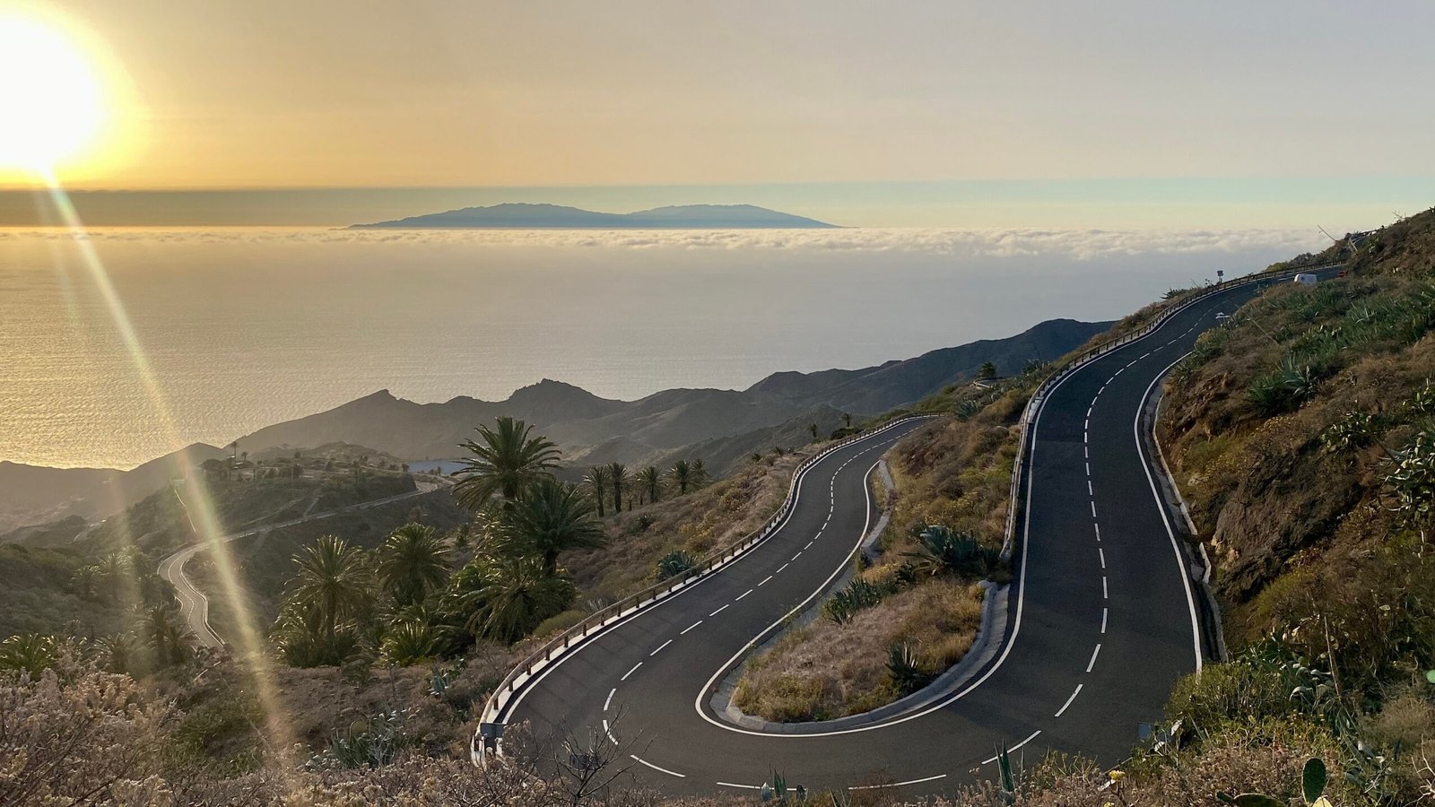 A winding road leads to a sunset over the ocean, casting warm hues across the sky and reflecting on the water.